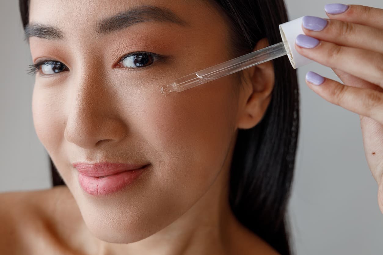 Closeup einer lächelnden, ostasiatischen Frau mit langen schwarzen Haaren, die mit einer Pipette Serum auf ihre Wange gibt